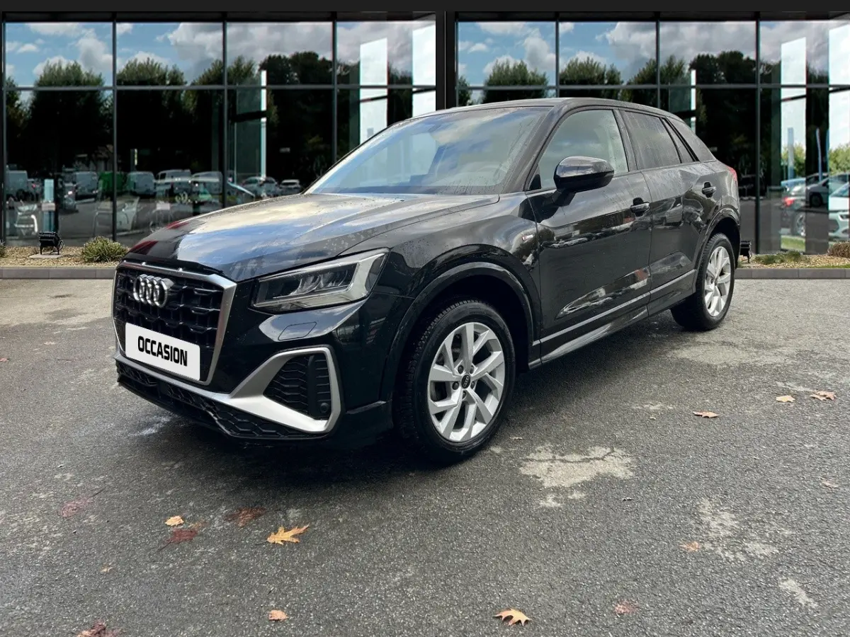 Audi Q2 noir vue 3/4 avant droit, avec calandre distinctive et jantes argentées sur parking devant vitres réfléchissantes.