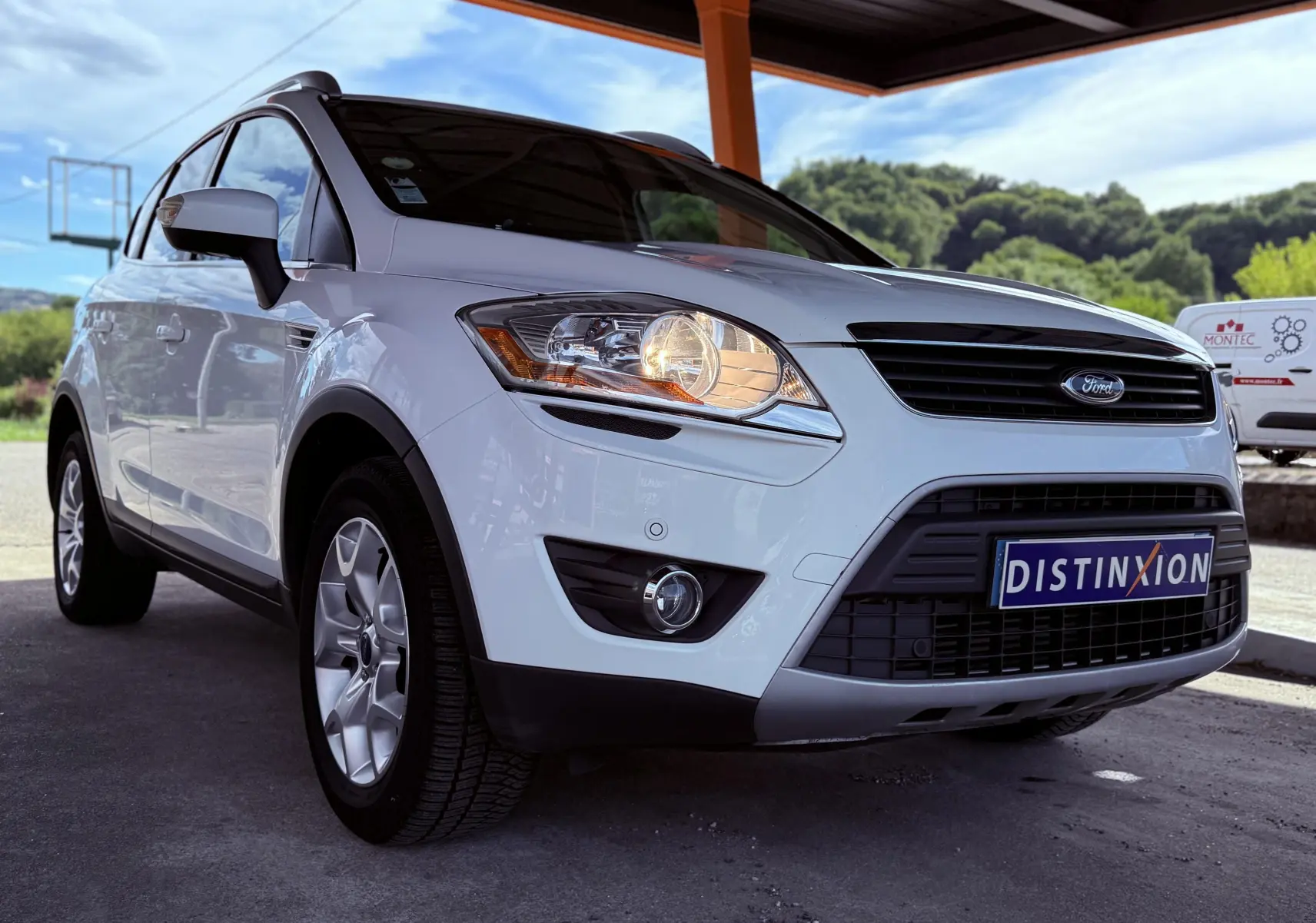 Ford Kuga blanc vu de face droite sous abri, phare avant allumé et calandre noire avec logo Ford.