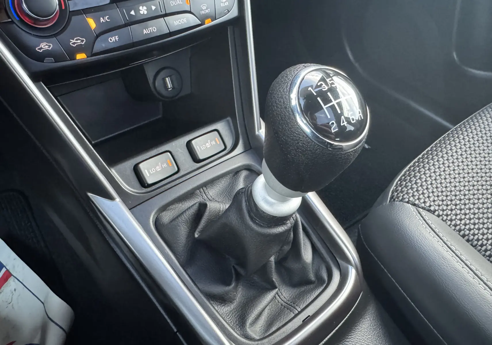 Gros plan sur le levier de vitesses manuel noir et argenté du Suzuki S-Cross blanc 2023 avec commandes de sièges chauffants visibles.