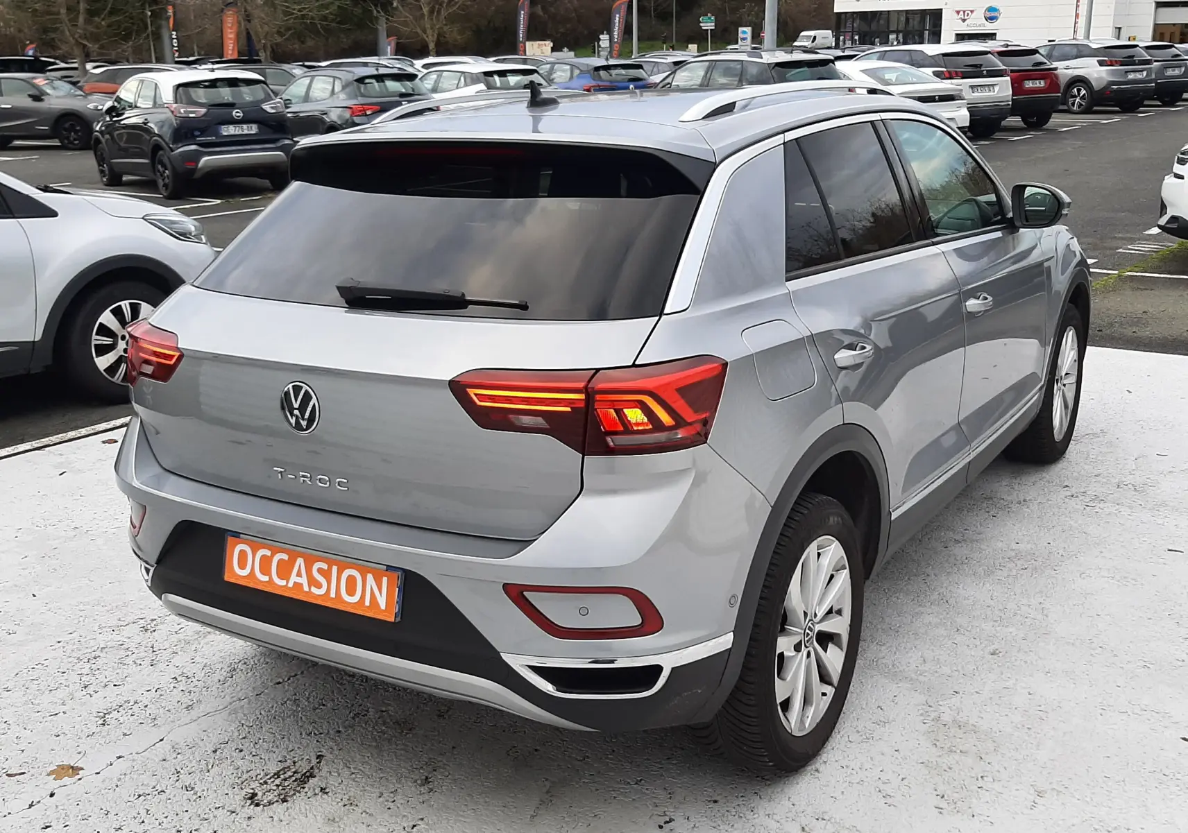 Volkswagen T-Roc gris pyrite vu de l'arrière droit, feux LED allumés, vitres arrière surteintées, sur parking extérieur.