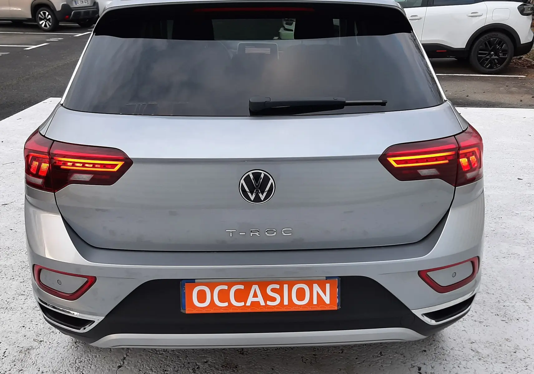 Vue arrière d'un Volkswagen T-Roc gris pyrite avec feux allumés et plaque orange "OCCASION" sur parking.