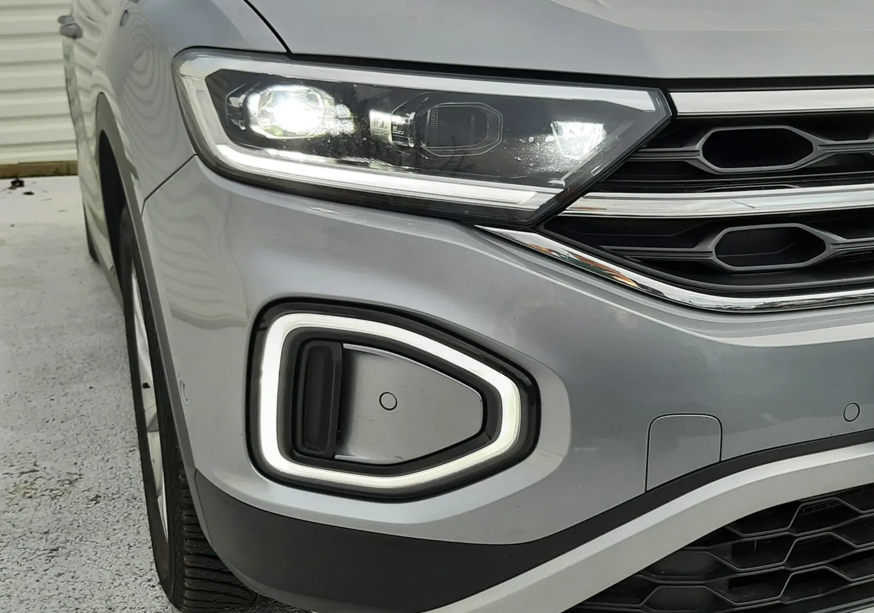 Gros plan sur l'avant gauche gris pyrite du Volkswagen T-Roc 2023 avec phare LED allumé et calandre distinctive.