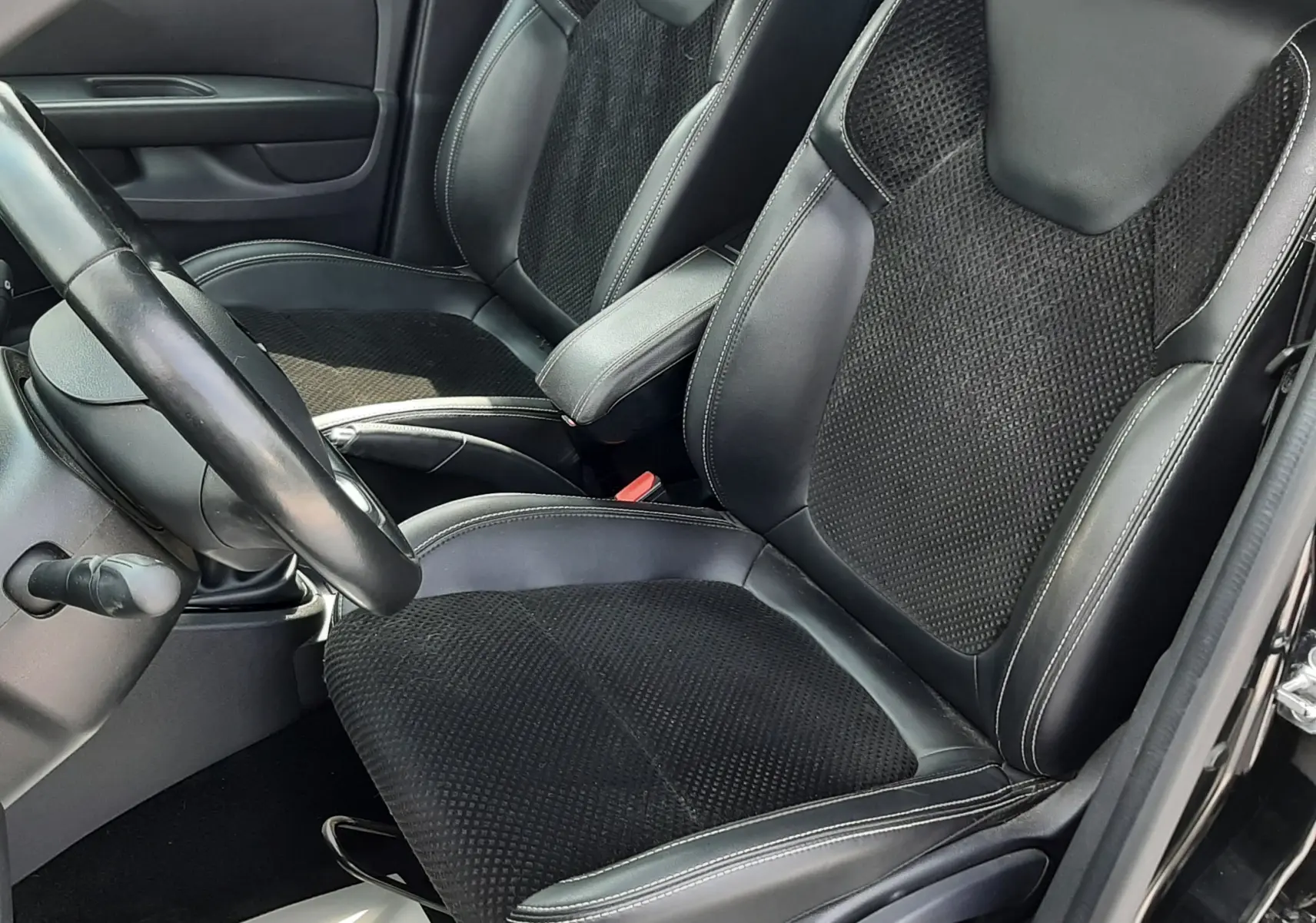Vue intérieure côté conducteur de la Renault Clio IV noire, mettant en valeur les sièges mixtes cuir et tissu noir avec surpiqûres blanches.