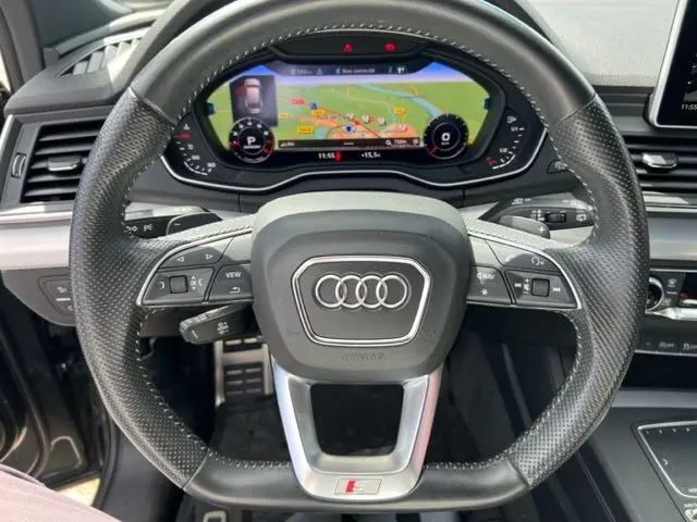 Vue rapprochée du volant en cuir perforé d'un Audi Q5 gris foncé 2018, avec tableau de bord numérique et affichage de navigation.
