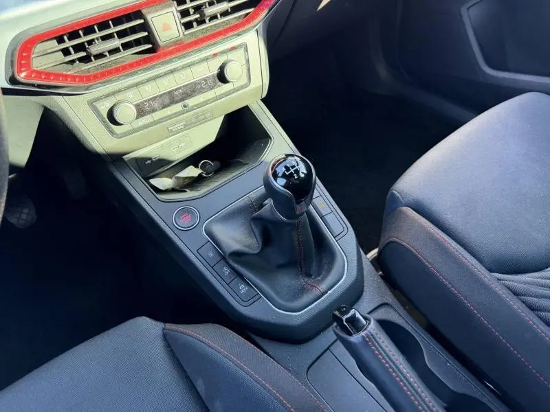 Vue intérieure centrée sur la console centrale d'une SEAT Ibiza 2025 blanche, avec levier de vitesse manuel et surpiqûres rouges.