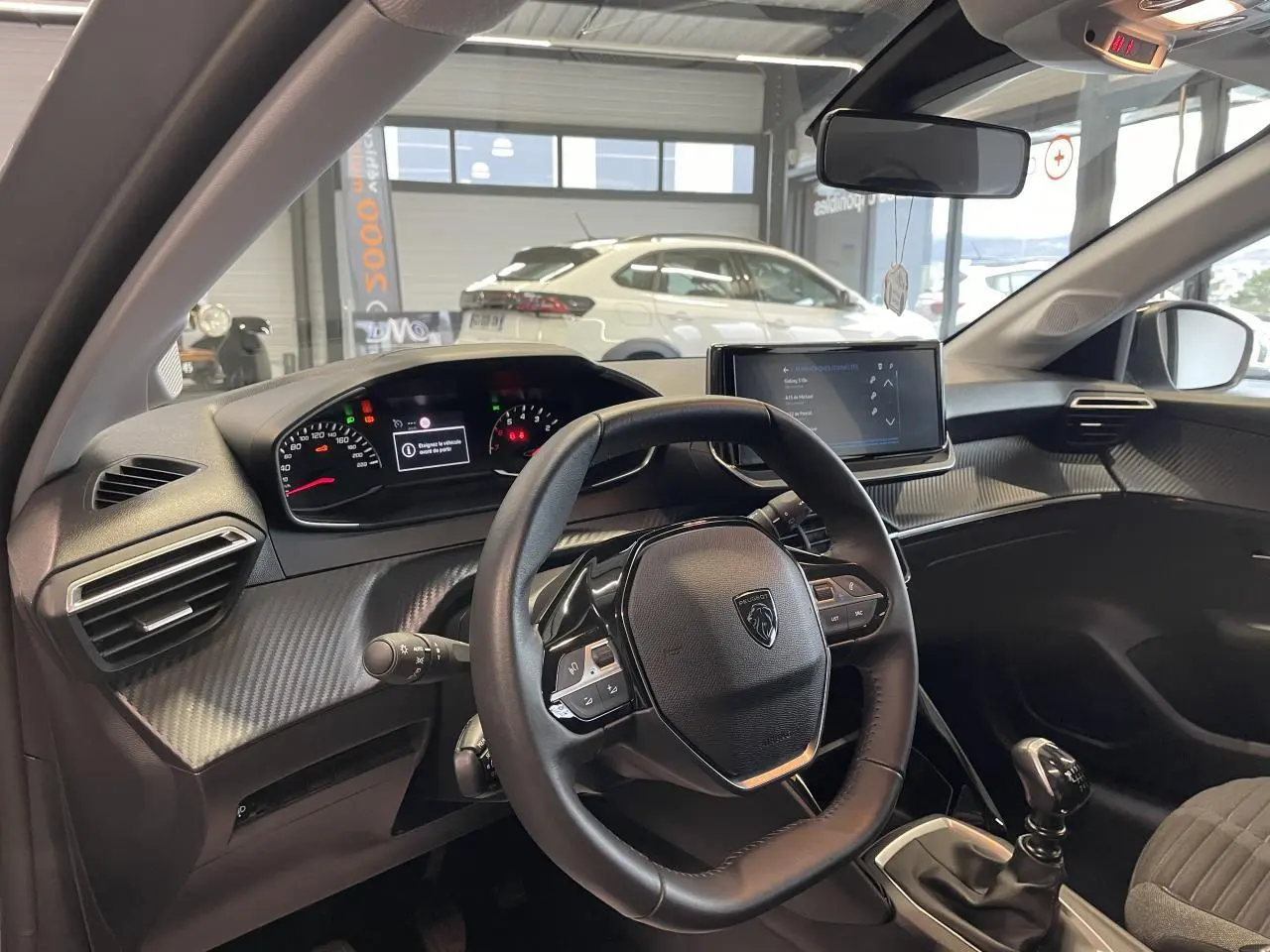 Vue intérieure du poste de conduite de la Peugeot 208 gris clair 2024, volant cuir et écran tactile central visible.