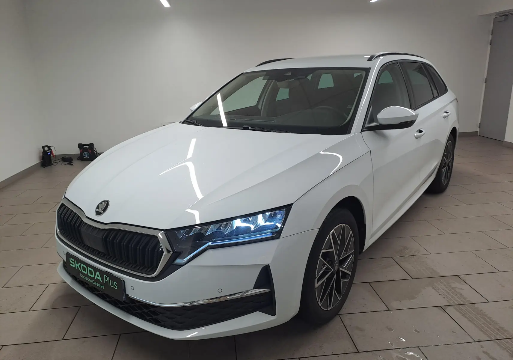 Vue 3/4 avant droite d'une Skoda Octavia Combi 2025 blanc Moon avec jantes alliage noires polies et phares LED allumés.