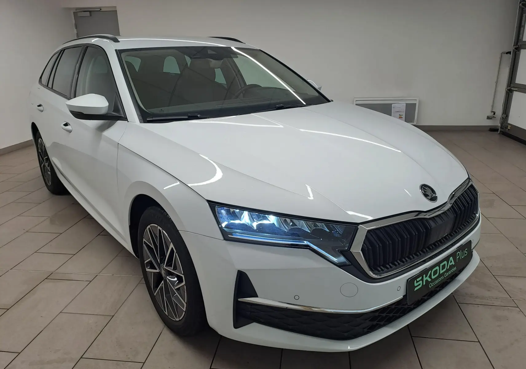 Vue 3/4 avant gauche d'une Skoda Octavia Combi blanche Moon avec phares allumés et jantes alliage noires polies.