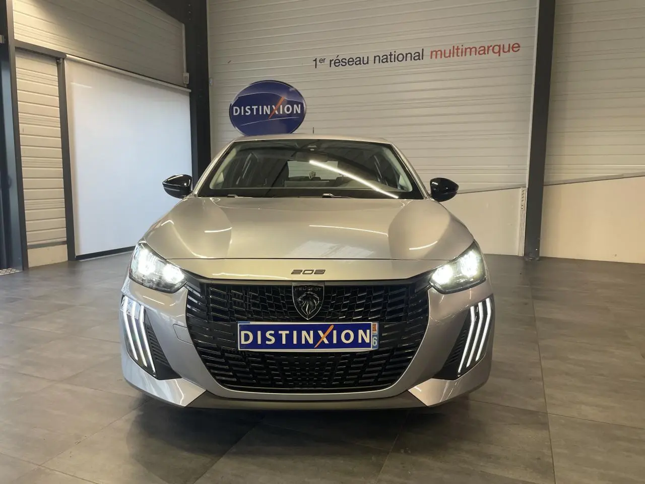 Vue frontale d'une Peugeot 208 gris clair 2024 avec calandre noire et feux LED allumés en intérieur showroom.