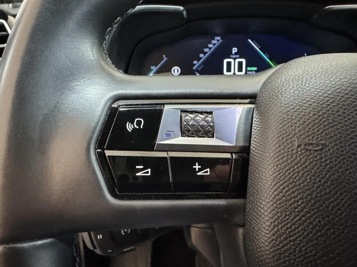 Gros plan sur les commandes gauche du volant cuir noir du DS7 Crossback E-TENSE 225 Bastille+ avec affichage digital en arrière-plan.