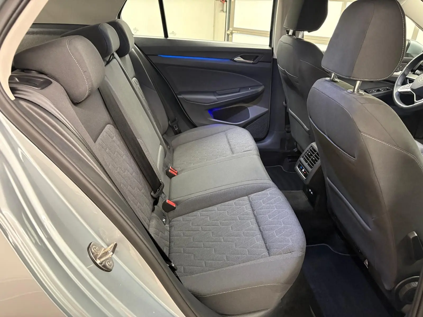 Vue intérieure côté arrière droit de la Volkswagen Golf 2.0 TDI 2025, sièges tissu gris avec éclairage d'ambiance bleu sur la porte.