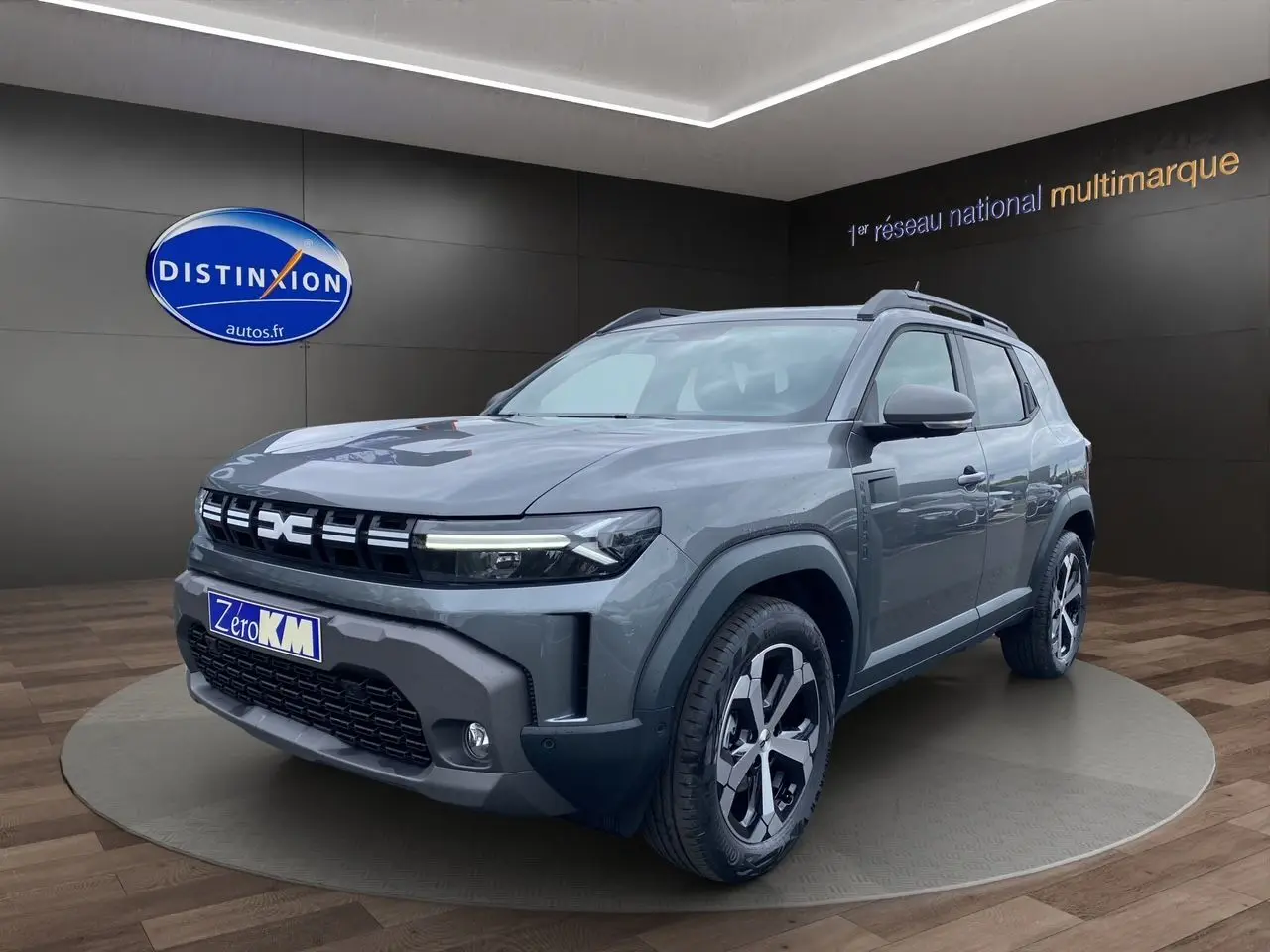 Vue 3/4 avant d'un Dacia Duster gris schiste hybride 2025 avec jantes noires et grises, sur podium showroom.