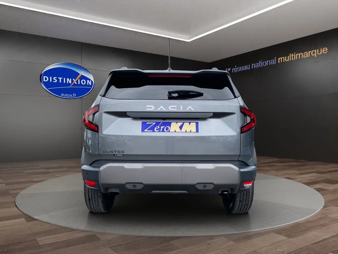 Vue arrière du Dacia Duster hybride gris schiste avec logo distinctif et plaque Zéro KM visible en showroom.