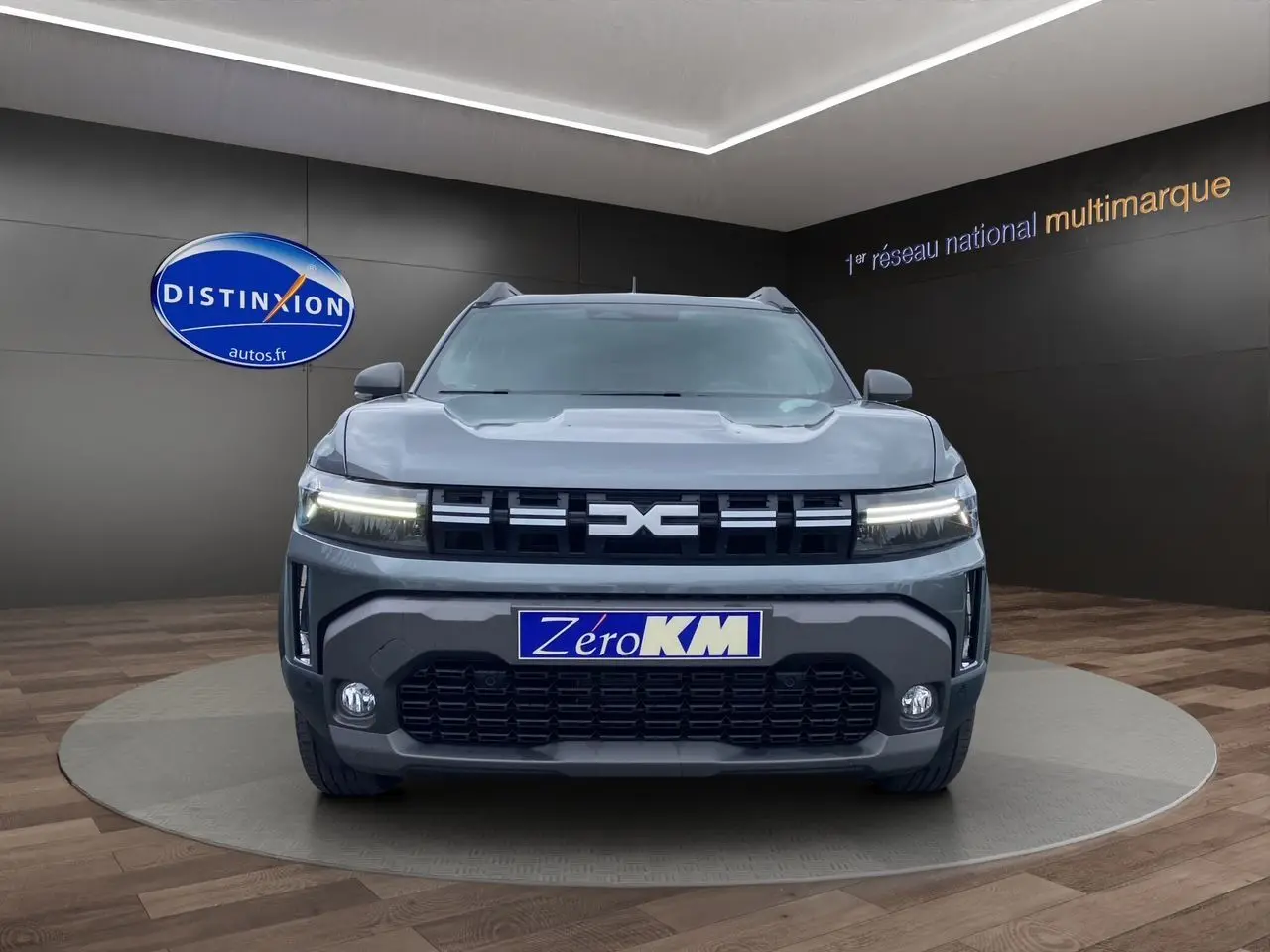 Vue frontale du Dacia Duster gris schiste 2025 avec calandre noire et phares LED allumés en showroom.