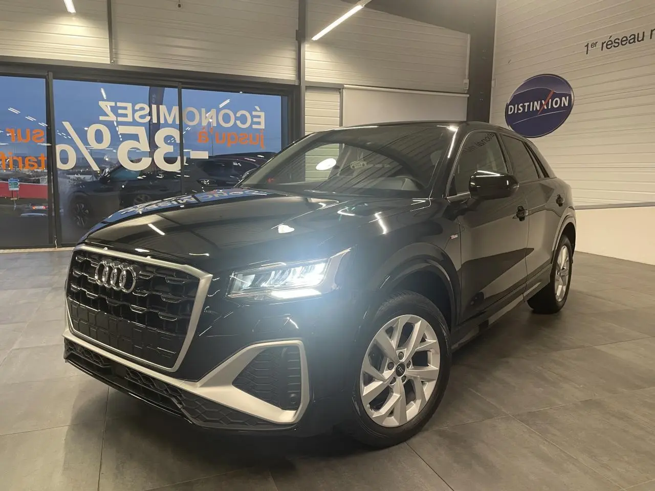 Audi Q2 noir métallisé en 3/4 avant droit avec phares allumés et jantes alliage 18 pouces style trapèze.