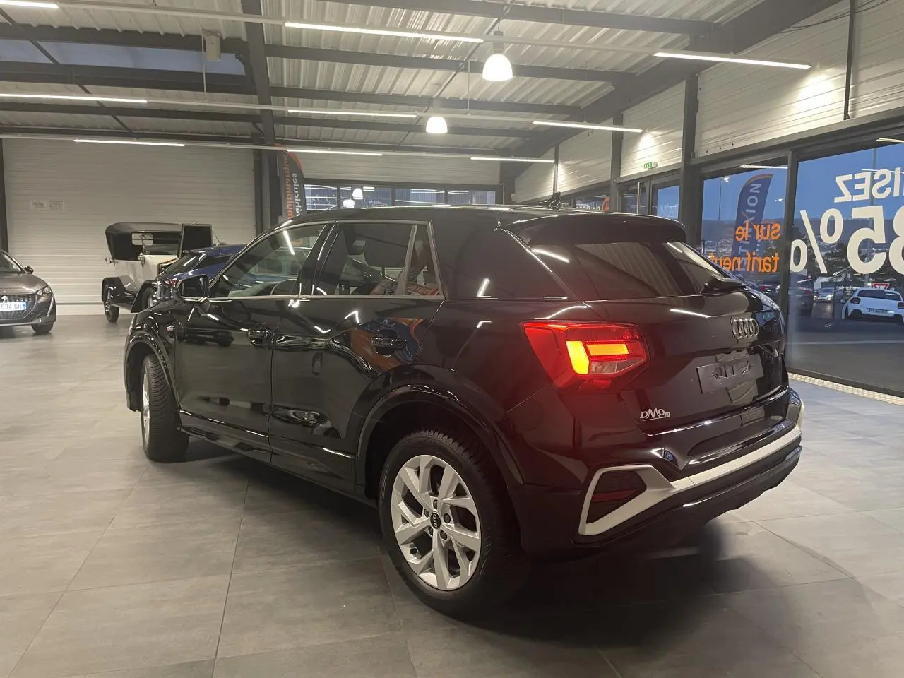 Audi Q2 noir métallisé vue 3/4 arrière droit en intérieur, feux arrière allumés et jantes en alliage visibles.