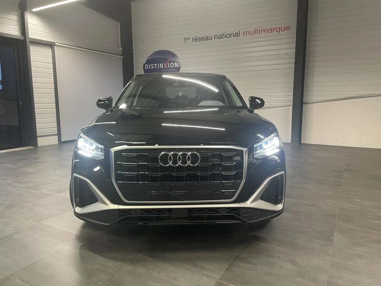 Audi Q2 S line noir métallisé vue de face avec phares Matrix LED allumés en intérieur showroom.