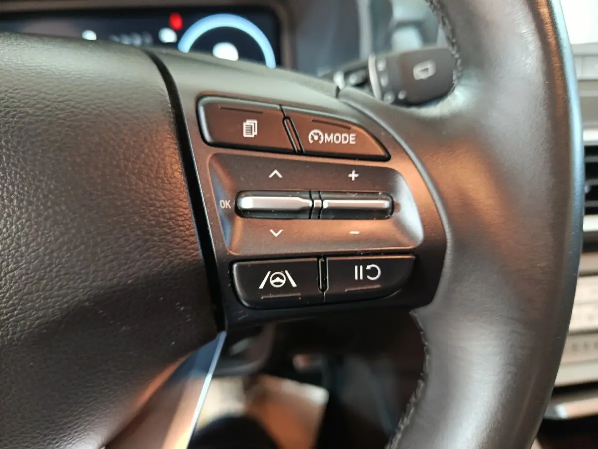 Gros plan sur les commandes du volant cuir noir du Hyundai Kona électrique bleu, avec boutons de régulateur et mode de conduite.