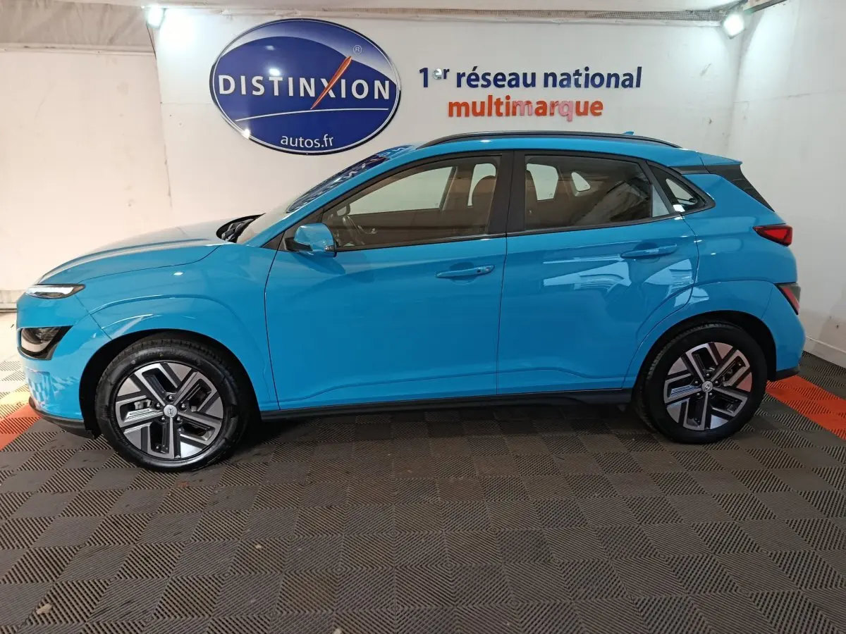 Vue de profil côté gauche d’un Hyundai Kona électrique bleu 2022 stationné en intérieur avec jantes spécifiques et rails de toit noirs.