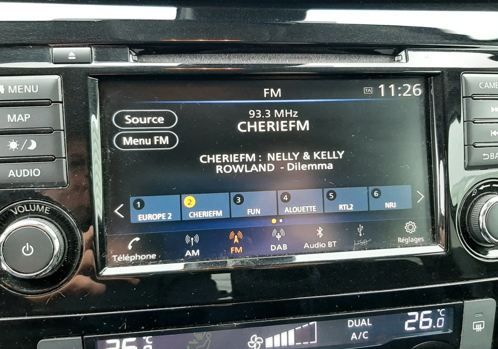 Vue rapprochée de l’écran multimédia et de la console de climatisation du Nissan Qashqai noir, affichant une radio FM et une température à 26°C.