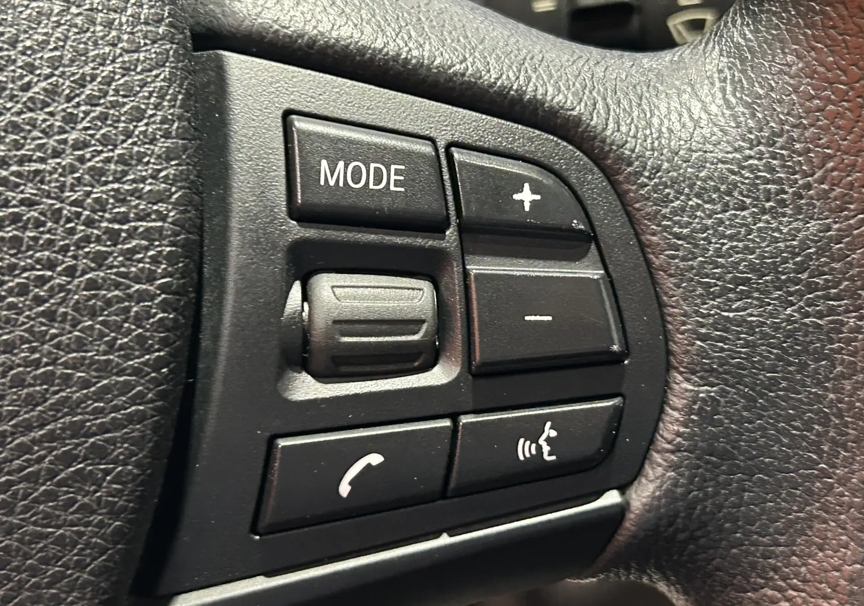 Gros plan sur les commandes noires du volant en cuir de la BMW Série 1 gris diesel, avec boutons mode et volume.