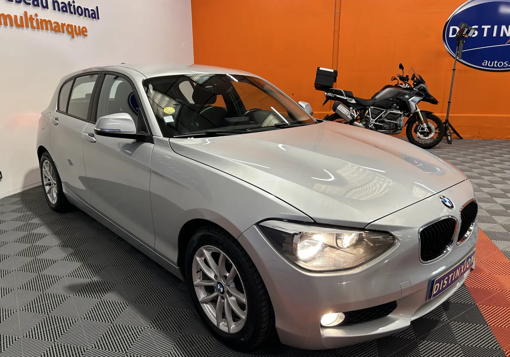 BMW Série 1 gris vue 3/4 avant droit avec phares allumés dans un showroom orange et blanc.