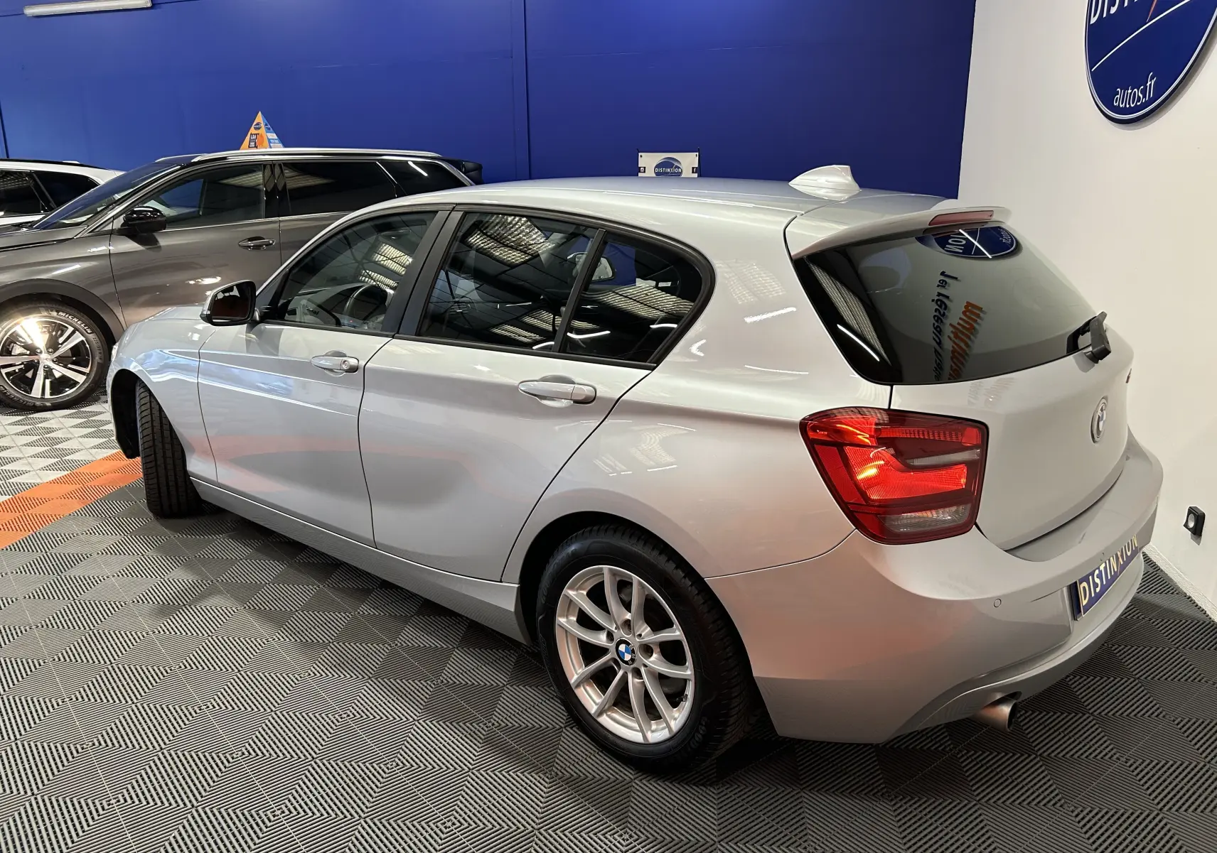 BMW Série 1 gris vue 3/4 arrière droit, compacte 5 portes avec jantes alliage et feux arrière allumés.