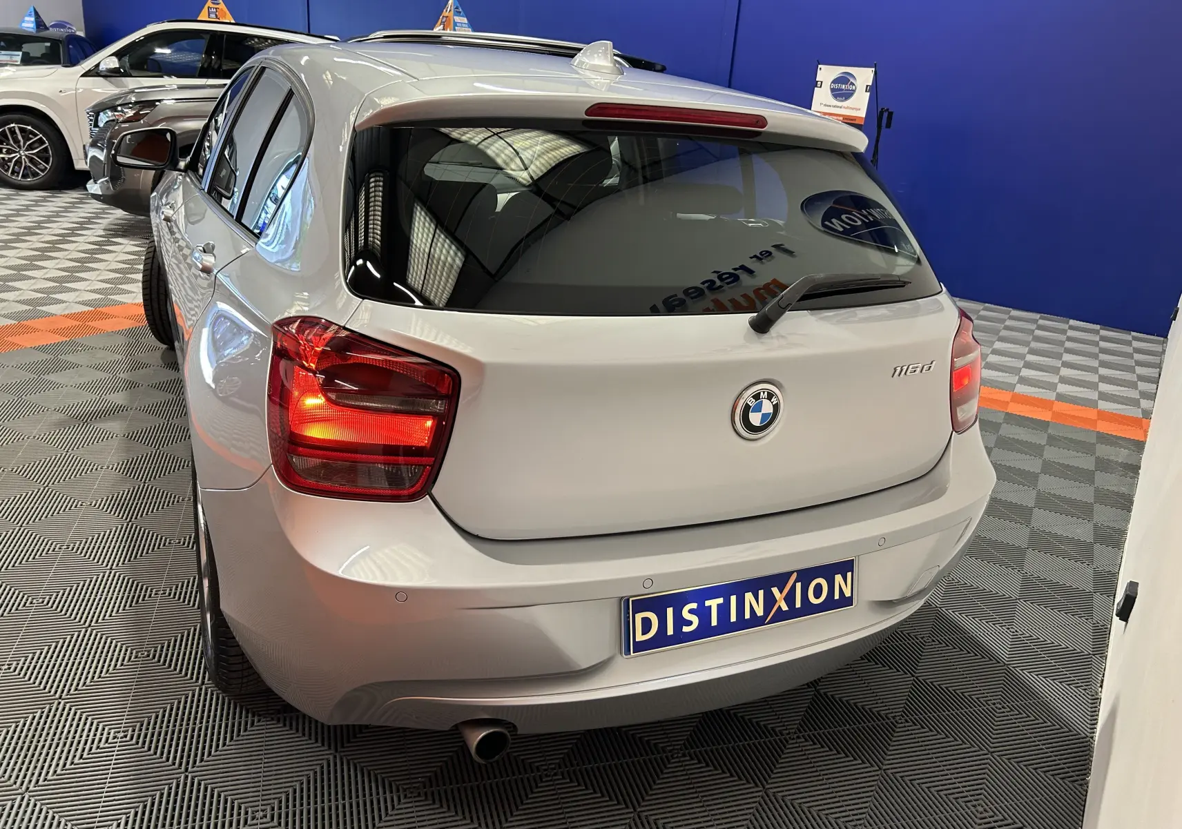 Vue arrière droite d'une BMW Série 1 gris clair 116d avec feux allumés dans un showroom.