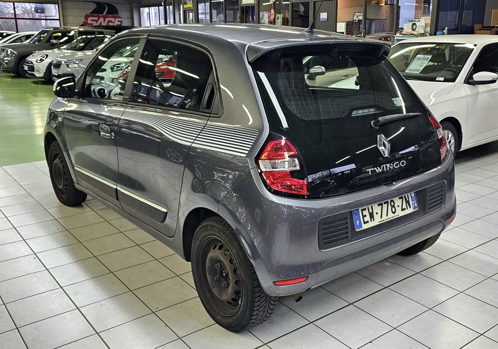 Vue 3/4 arrière droite d'une Renault Twingo III gris lunaire 2018 en intérieur, avec jantes acier noires et bandes blanches latérales.