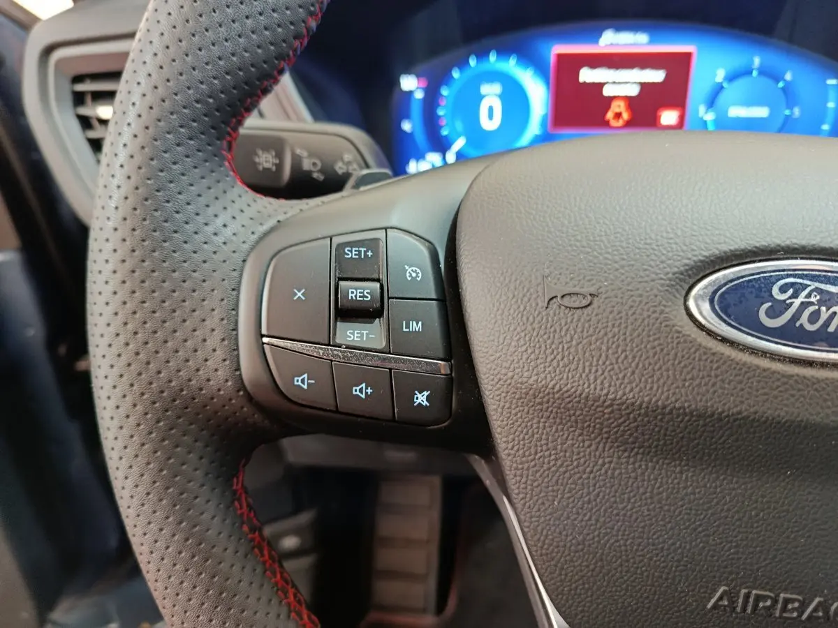 Gros plan sur le volant cuir noir perforé avec commandes multifonctions du Ford Kuga 2024, tableau de bord numérique bleu en arrière-plan.