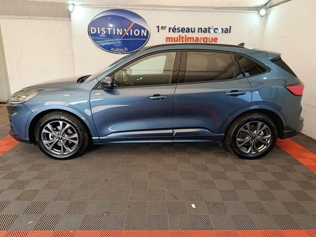 Vue latérale droite d’un Ford Kuga 2024 bleu métallisé St Line avec jantes alliage et vitres teintées, en intérieur showroom.