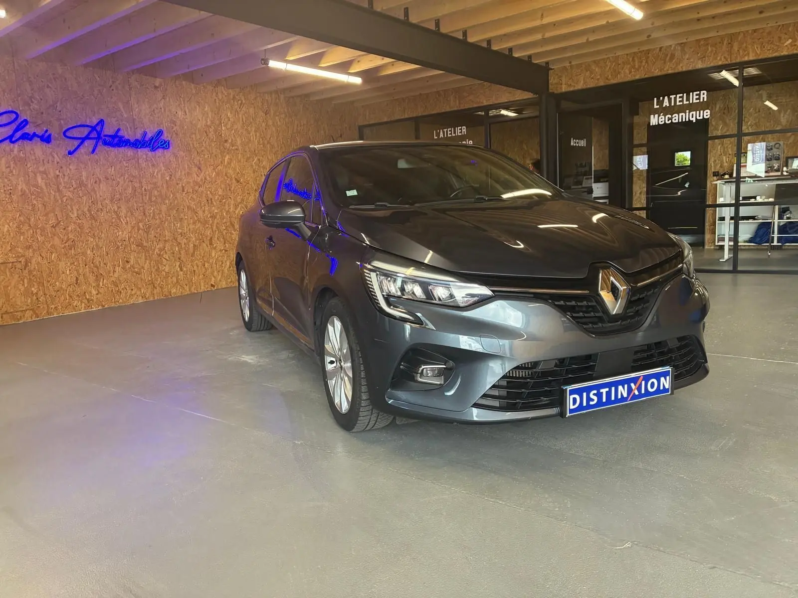 Renault Clio gris 3/4 avant droit dans un showroom avec éclairage intérieur et logo bleu au mur.