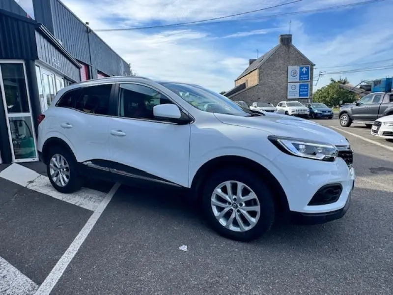 Renault Kadjar blanc en vue 3/4 avant droit, avec jantes alliage et barres de toit visibles.