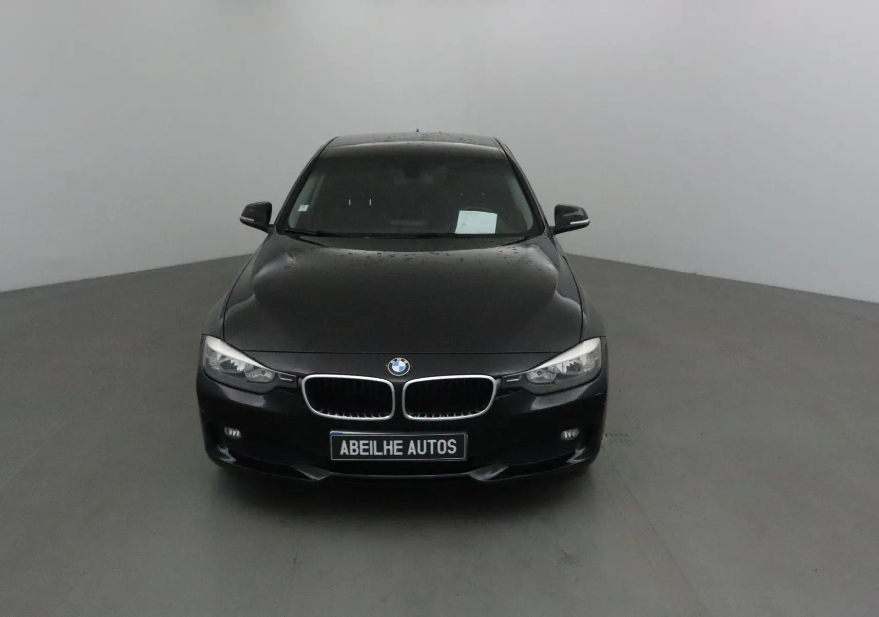 BMW Série 3 316D noire vue de face dans un studio, avec calandre caractéristique et plaque Abeilhe Autos visible.