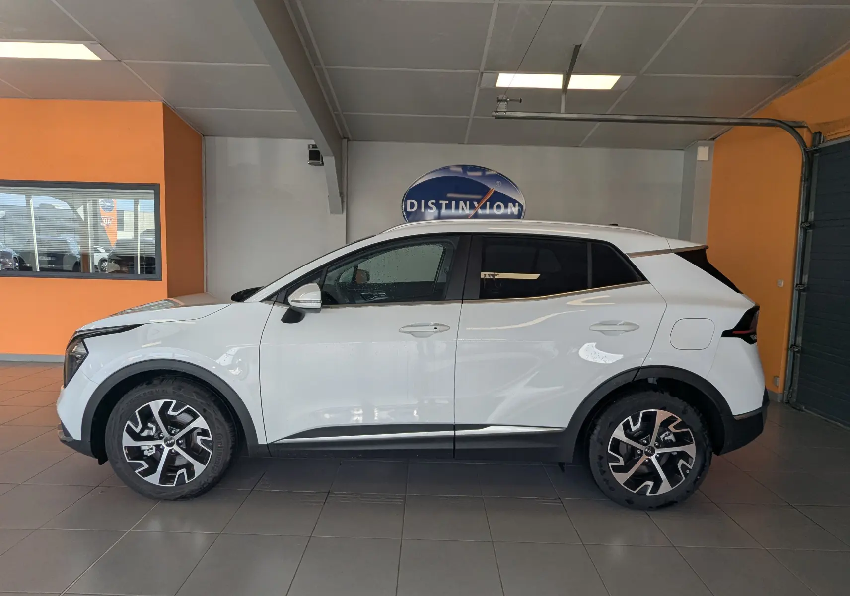 Vue de profil côté gauche d'un KIA Sportage blanc 2025 avec jantes alliage bicolores et vitres teintées, en intérieur showroom.