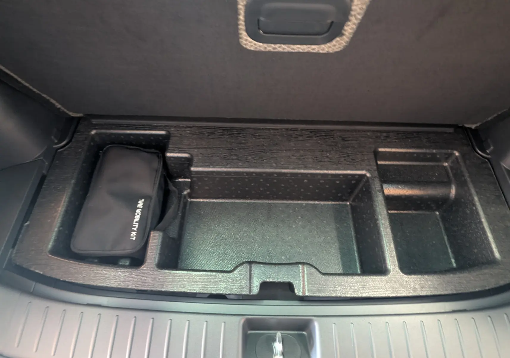 Coffre ouvert du Kia Sportage blanc 2025, vue plongeante sur compartiment de rangement avec trousse de mobilité noire.