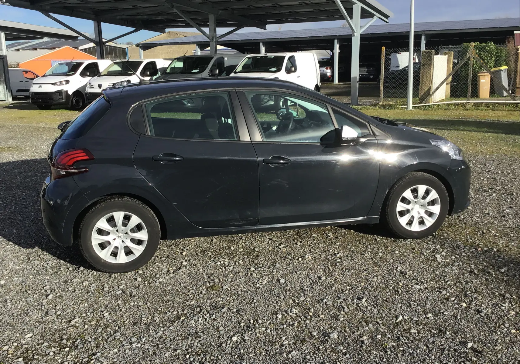 Peugeot 208 noire vue de profil côté gauche, garée sur un sol gravillonné avec des véhicules utilitaires en arrière-plan.