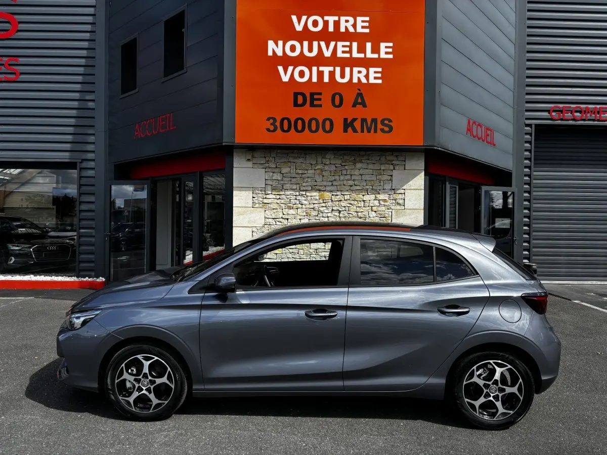 Vue latérale gauche d'une MG3 gris clair 2025 garée devant un bâtiment avec panneau orange "Votre nouvelle voiture".