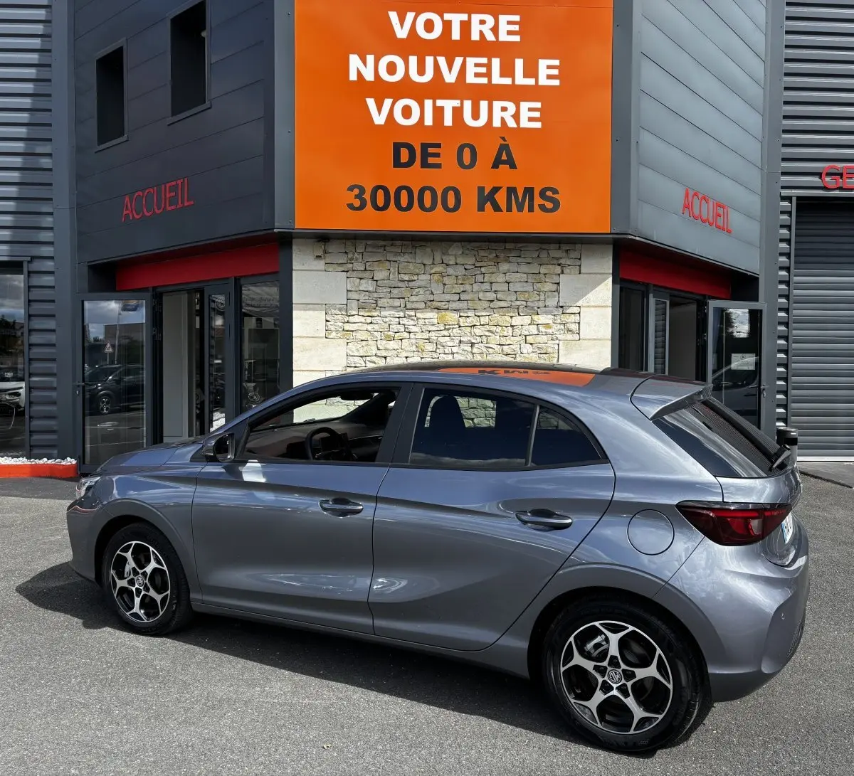 Vue latérale droite d'une MG3 gris clair garée devant un bâtiment avec panneau orange "Votre nouvelle voiture de 0 à 30000 kms".
