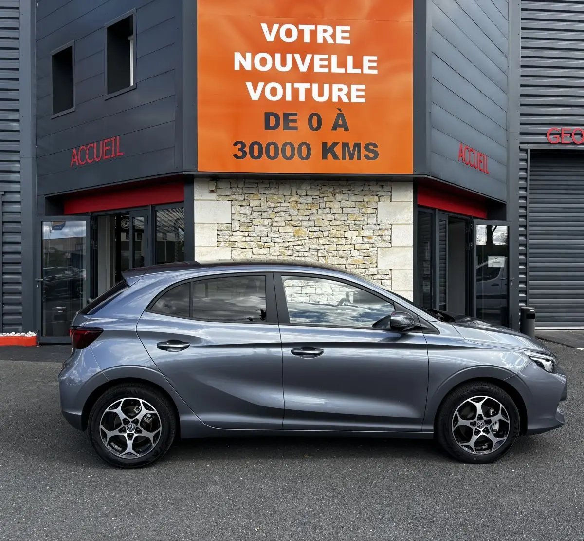 Vue de profil côté gauche d'une MG3 gris clair 2025 garée devant un bâtiment avec un panneau orange "Votre nouvelle voiture".