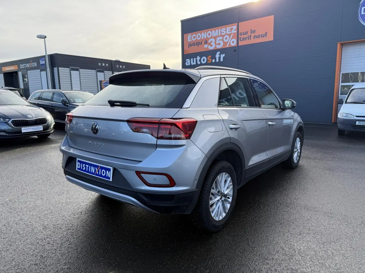 Volkswagen T-Roc gris pyrite vu en 3/4 arrière droit sur parking devant un bâtiment commercial.