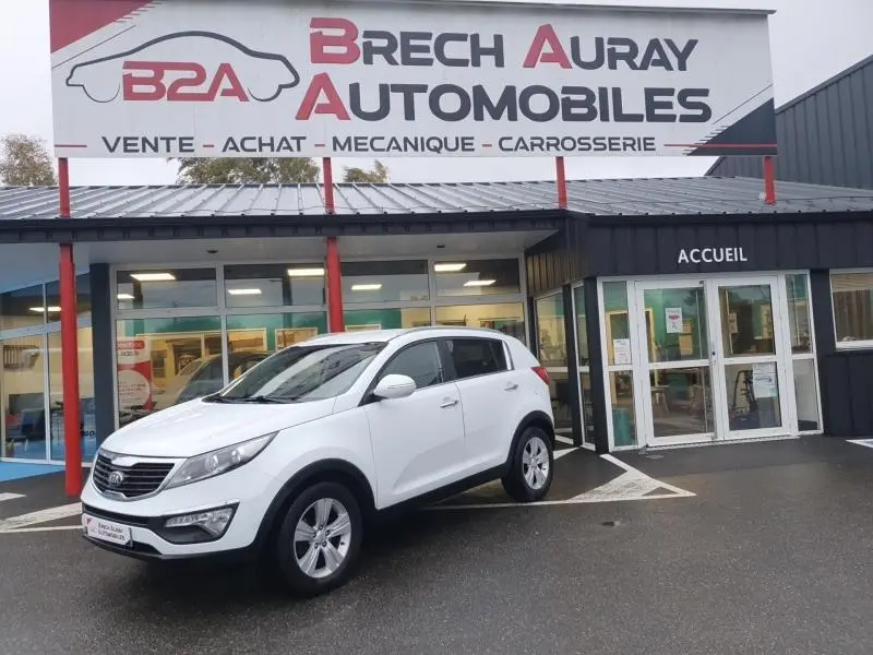 KIA Sportage blanc en 3/4 avant droit stationné devant un garage avec enseigne Brech Auray Automobiles.