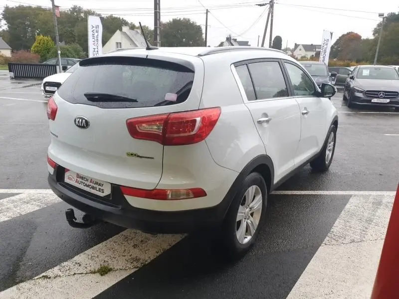 KIA Sportage blanc vu en 3/4 arrière droit, avec barres de toit et attelage visible sur parking humide.