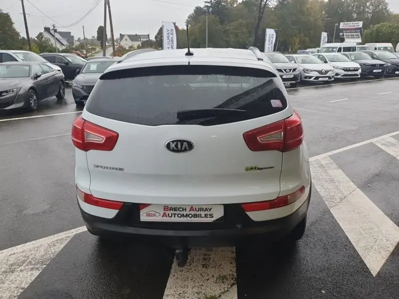 Vue arrière d'un KIA SPORTAGE blanc de 2013 avec feux rouges et logo KIA bien visibles sur un parking humide.