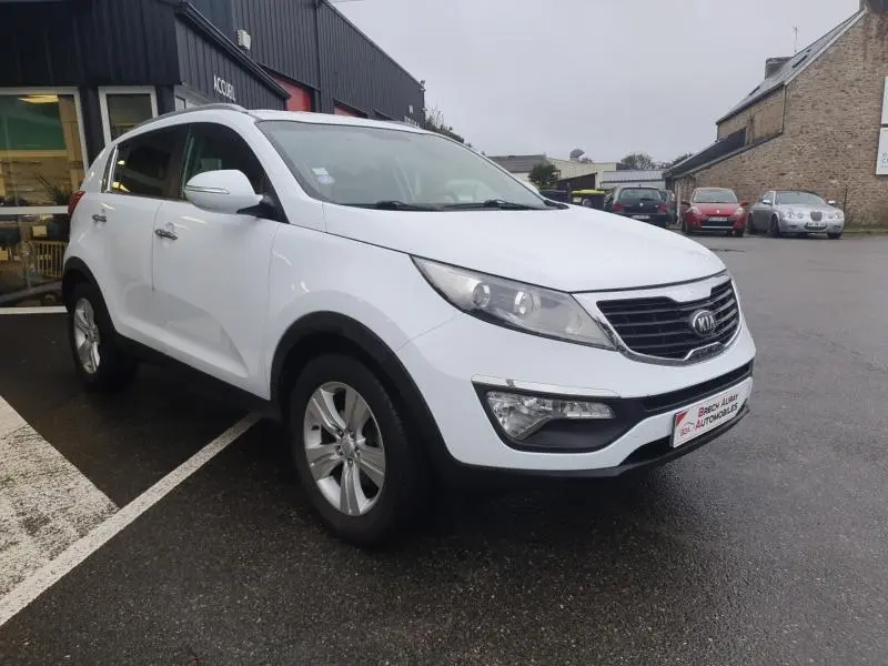 KIA Sportage blanc vu en 3/4 avant droit, avec calandre chromée et jantes alliage sur parking humide.