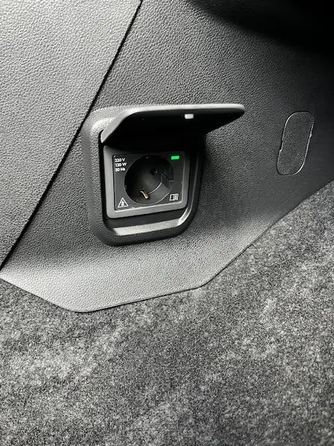 Prise 220V intégrée dans le coffre du Peugeot Rifter gris foncé, avec trappe de protection ouverte.