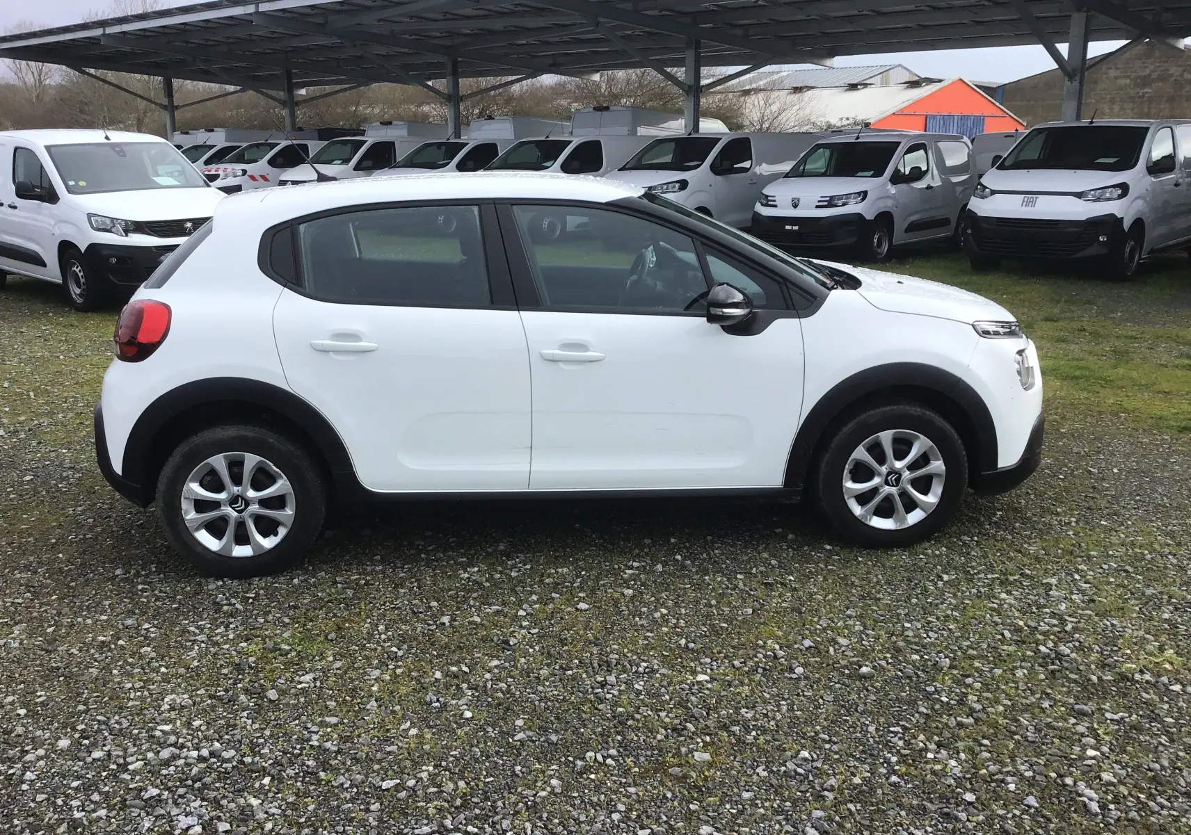 Citroën C3 blanc vue de profil côté gauche sur un parking avec plusieurs véhicules utilitaires en arrière-plan.