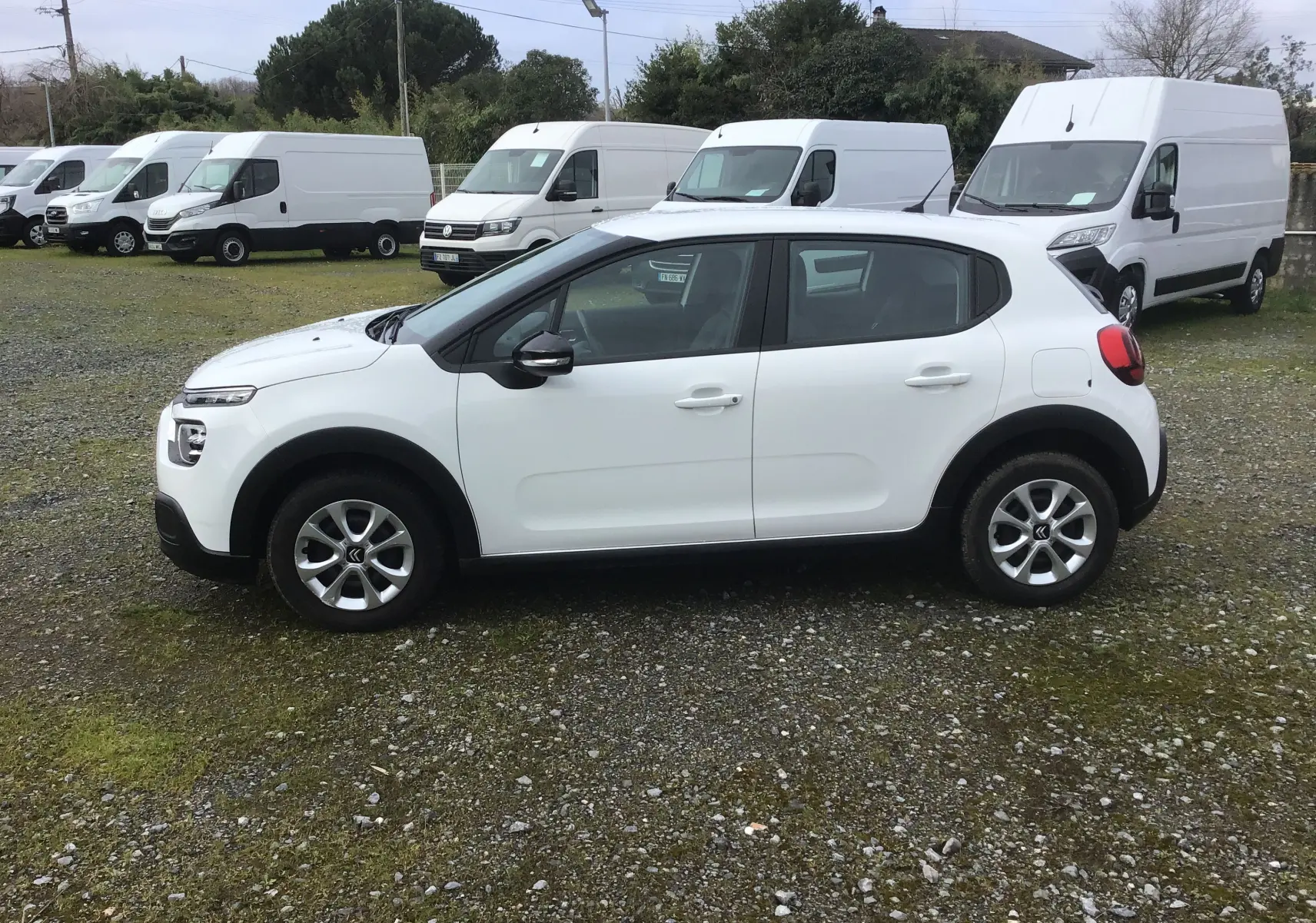 Citroën C3 blanc vue de profil côté gauche, avec jantes alliage et entourée de fourgons blancs en arrière-plan.