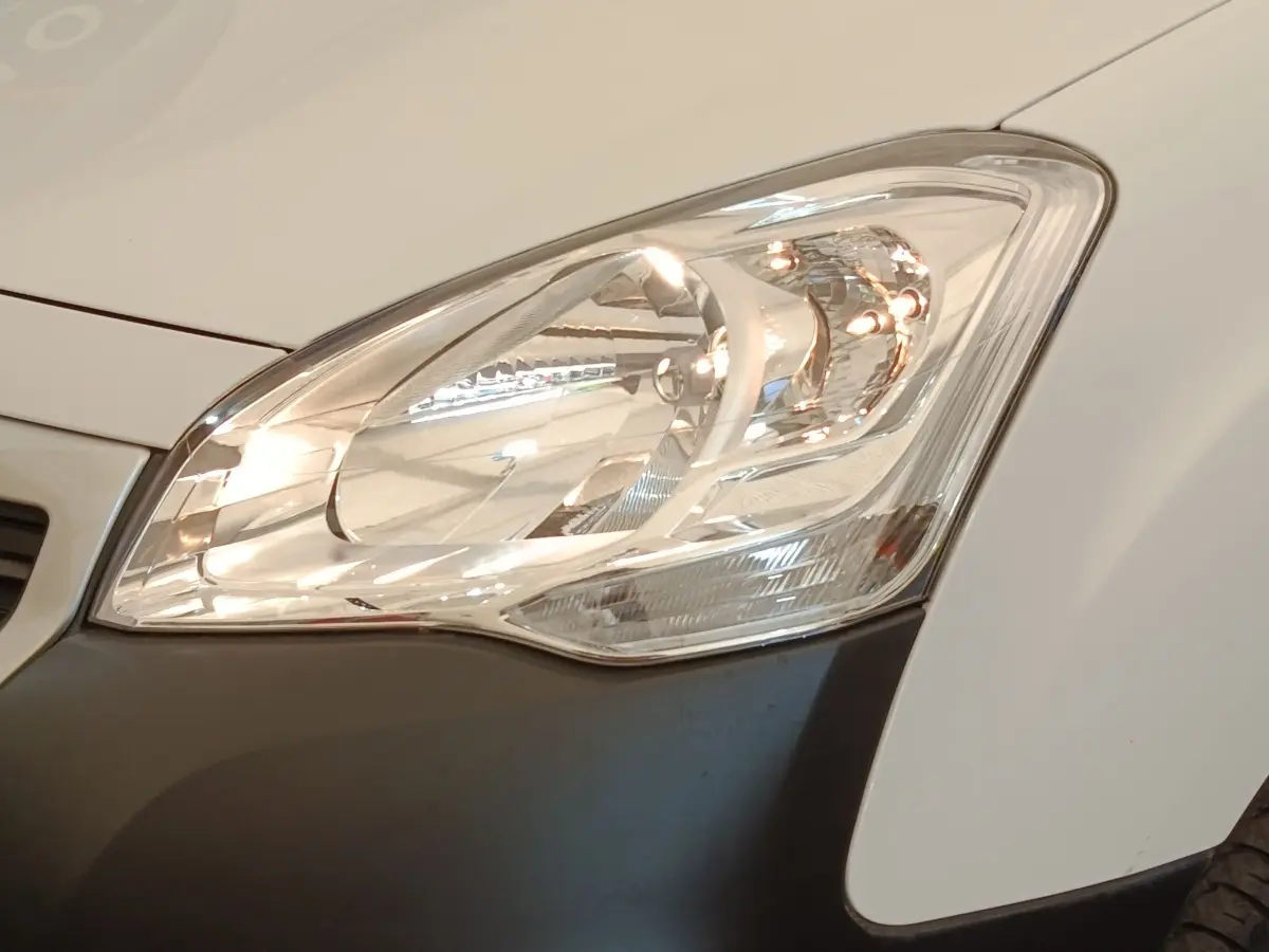 Gros plan sur le phare avant droit du Peugeot Partner blanc, avec partie inférieure du pare-chocs noir visible.