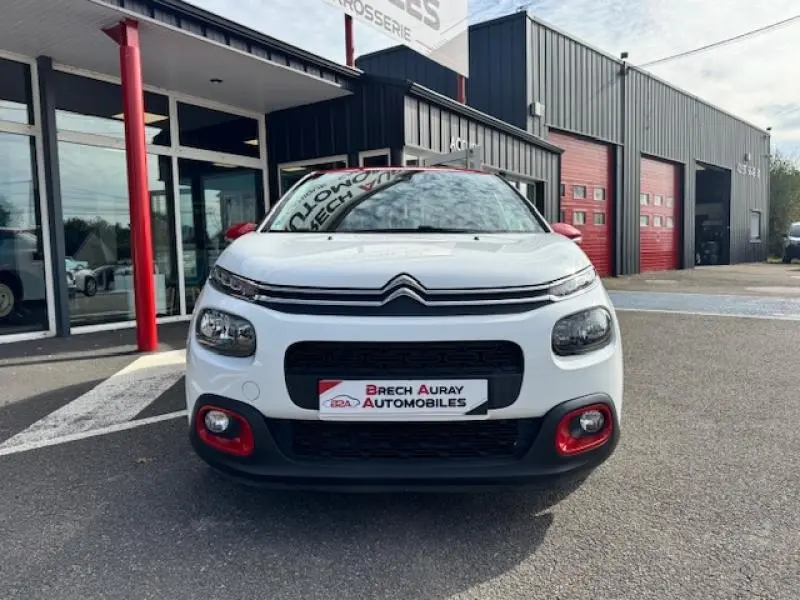Vue avant d'une Citroën C3 2017 blanc banquise avec détails rouges sur pare-chocs et rétroviseurs, devant un garage.